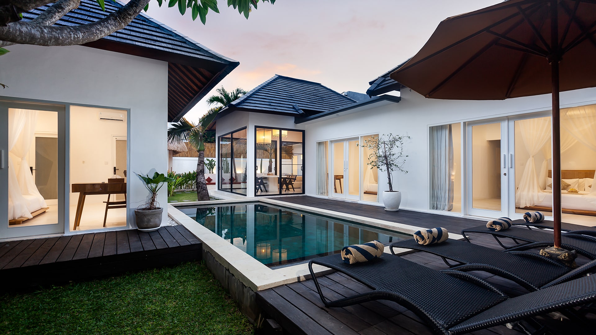 Villa courtyard at dusk with pool, timber deck, and lit bedrooms on either side
